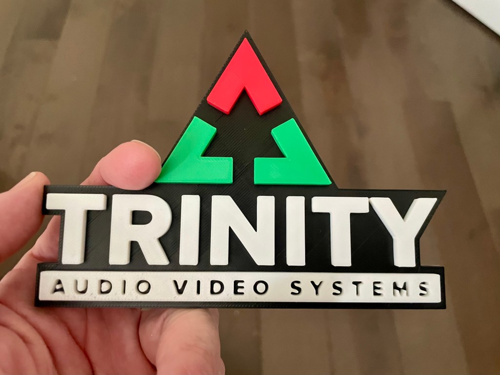 A logo held up in front of a brown floor. The logo has stylized triangular elements and says "TRINITY AUDIO VIDEO SYSTEMS".
